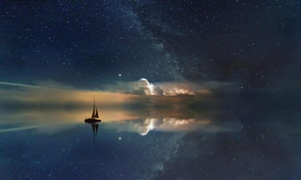 Lone sailboat floating on calm water beneath a star‑filled night sky and glowing Milky Way.
